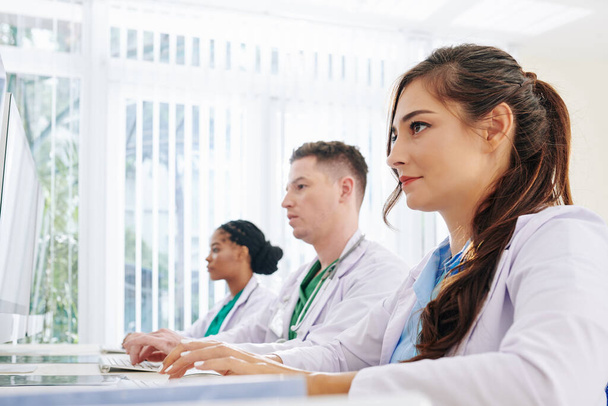Серьезные молодые врачи, работающие за компьютерами в офисе, отвечая на вопросы пациентов в Интернете и заполняя документы - Фото, изображение