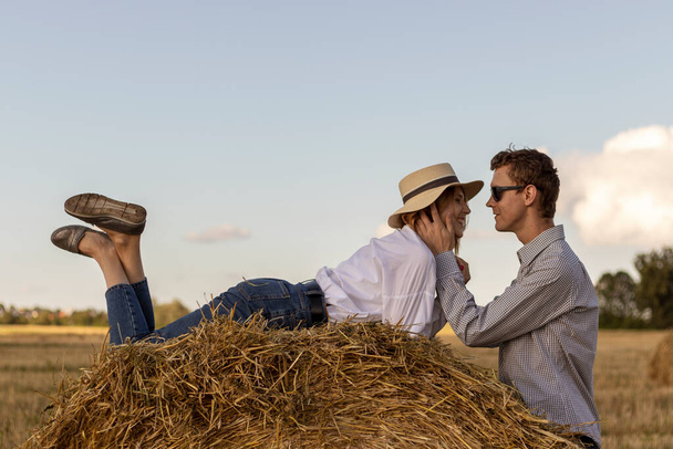 Портрет закоханої пари в полі в сільській місцевості. Молода жінка в блакитних джинсах, сорочка і солом'яний капелюх лежить на стовбурі сіна. Поруч з дівчиною її хлопець
. - Фото, зображення