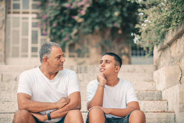 szczęśliwy nastolatek młody chłopiec odpoczynek na schodach rozmawiać ze starszym ojcem cieszyć wypoczynek rodzinny weekend. - Zdjęcie, obraz