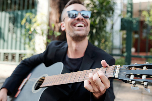 Güneş gözlüklü neşeli müzisyen gitar çalıyor, seçici odaklanıyor. - Fotoğraf, Görsel