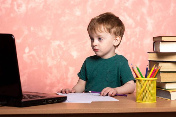 ένα μικρό παιδί κοιτάζει σοβαρά σε ένα σημειωματάριο με ένα χρωματιστό μολύβι στο χέρι του. μαθήματα ζωγραφικής στο διαδίκτυο. εξ αποστάσεως εκπαίδευση. - Φωτογραφία, εικόνα