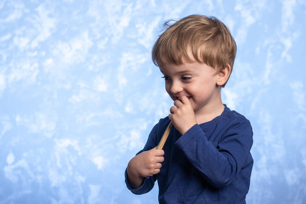 Ein kleiner Junge mit einem Lächeln putzt seine Zähne mit einer Bambuszahnbürste auf blauem Hintergrund. horizontales Foto. Platz für Text. Umweltfreundliche Morgenhygiene für ein Kind - Foto, Bild