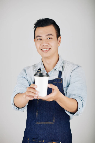 Улыбающийся владелец кофешопа в джинсовом фартуке дает чашку кофе - Фото, изображение