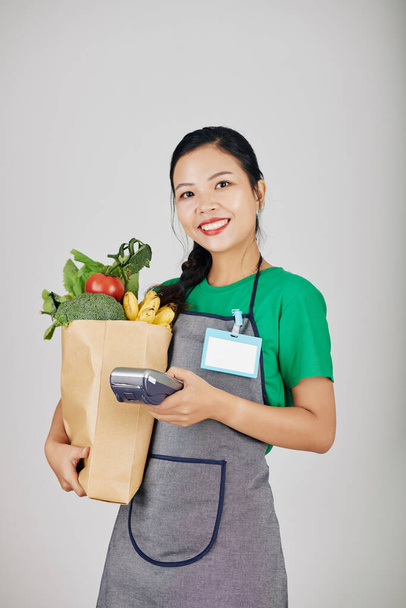 Porträt eines lächelnden jungen vietnamesischen Supermarktarbeiters mit Papierpaket mit frischem Gemüse und Zahlungsterminal - Foto, Bild