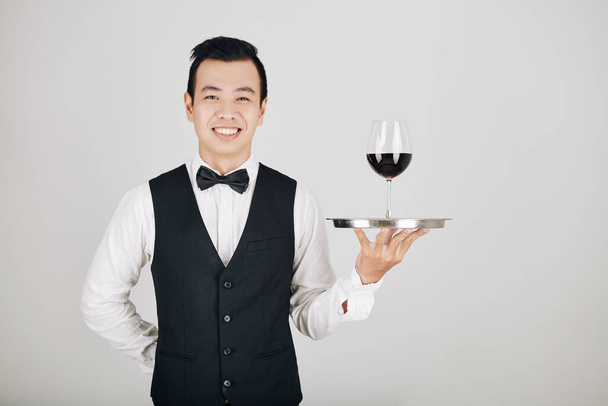Retrato del joven camarero asiático sonriente sosteniendo bandeja con copa de vino tinto
 - Foto, imagen