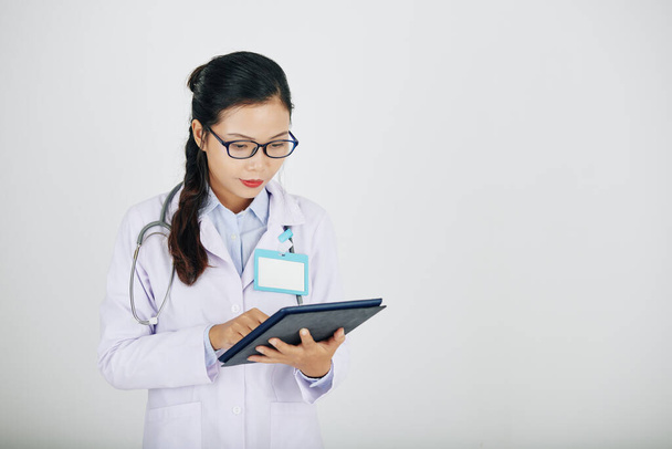 Junge asiatische Medizinpraktikantin überprüft Informationen auf Tablet-Computer, isoliert auf weiß - Foto, Bild