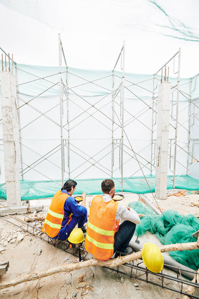 Budowniczowie w jasnych kamizelkach jedzący lunch na placu budowy, widok z tyłu - Zdjęcie, obraz