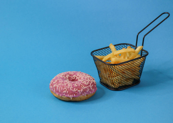 Um donut rosa e batatas fritas em uma cesta preta de ferro à direita em um fundo azul e com espaço para texto à esquerda. Vista lateral em close-up
. - Foto, Imagem
