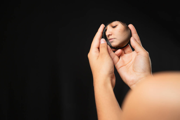 Hispanic woman looking at herself in a mirror with black background - close up hands holding a small mirror with the reflection of a face in it - reflection of her face - 写真・画像