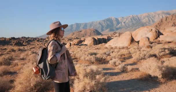 Sorrindo jovem mulher na moda em chapéu andando pela paisagem do deserto ao nascer do sol
 - Filmagem, Vídeo