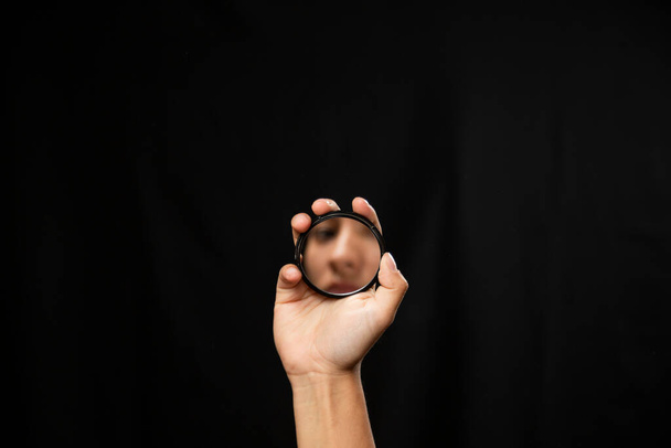 Hispanic woman looking at the mirror with black background - hand holding a small mirror with the reflection of a face in it - hand focused - Photo, Image