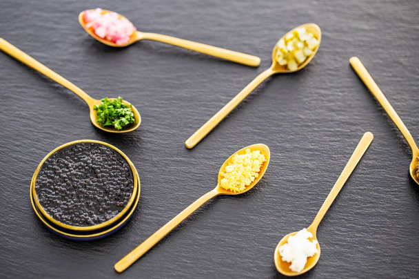 Caviar de esturjão preto real de alta qualidade em uma lata dourada de metal pode deixar de lado colheres douradas com picles, cebolas vermelhas, ovos e ervas em um fundo de ardósia. Vista lateral
. - Foto, Imagem