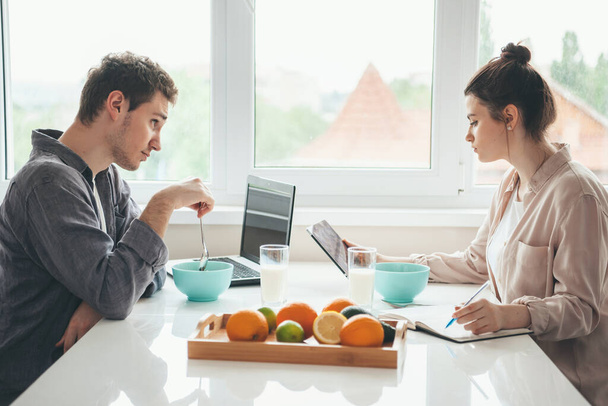 Homem caucasiano comendo cereais e usando um laptop está olhando para sua esposa beber leite e escrever algo
 - Foto, Imagem