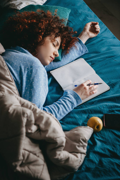 Felső nézet fotó egy göndör hajú nő kék pizsamában elalszik, miután megírta a házi feladatot a másolókönyvben - Fotó, kép