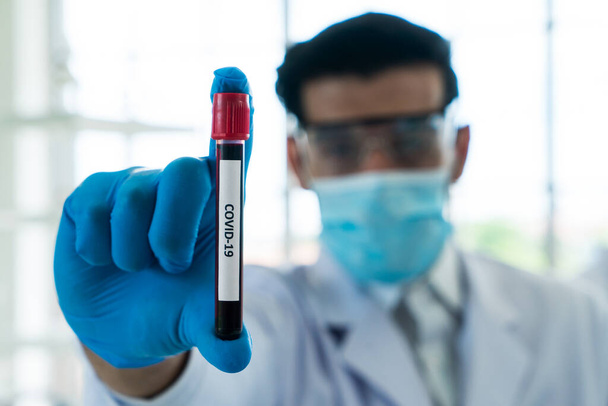 De wetenschapper draagt Masker en bril met Covid 19 bloedmonster in het laboratorium.. - Foto, afbeelding