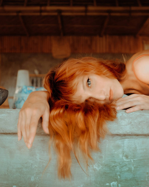 Attractive red hair model laying on kitchen countertops in Balinese Bungalow. Contemporary portraits looking through camera. - Photo, image