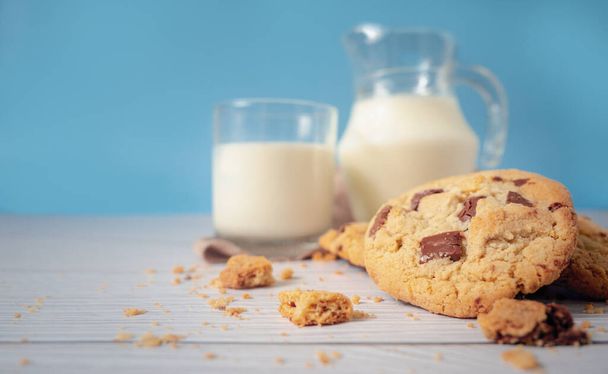 Lahodné domácí čokoládové sušenky, spojené s čerstvým mlékem ve sklenici a džbánem, položené na bílou dřevěnou podlahu a modrý ubrus na pozadí. Ráno pít pro zdraví a silné kosti. - Fotografie, Obrázek