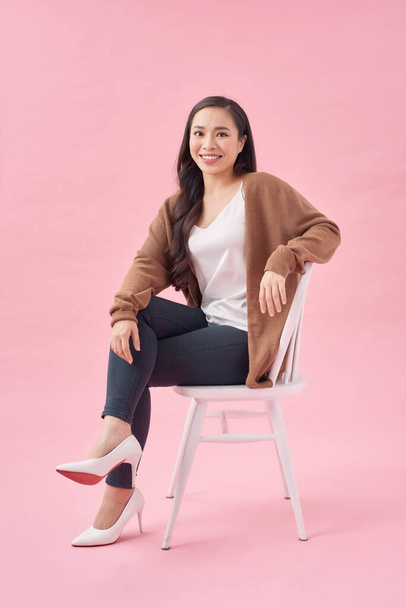 Portrait of a cheerful beautiful girl sitting on a chair and looking away isolated over pink background - Foto, immagini