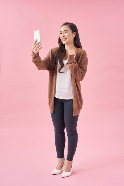 Retrato de una atractiva joven asiática usando teléfono móvil mientras está de pie con espacio de copia sobre fondo rosa
 - Foto, imagen