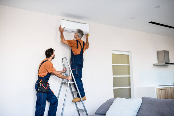 Exceptional service. Two workers in uniform, air conditioning masters using ladder while installing a new air conditioner in the apartment. Construction, maintenance and repair concept - Fotó, kép