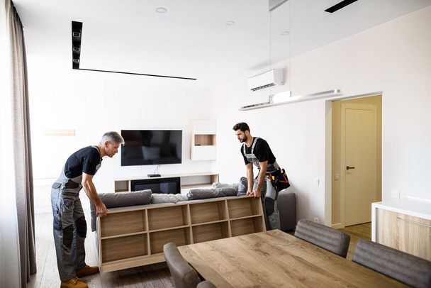 Moving with care. Full length shot of two contractor employees, young and aged worker in uniform moving personal belongings, furniture inside apartment. Moving service concept - 写真・画像
