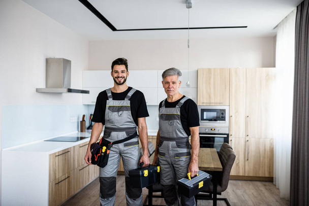 We make your life easier. Portrait of young and aged repairmen in uniform looking at camera with a smile, holding toolbox, ready for fixing kitchen hood. Repair service concept - 写真・画像