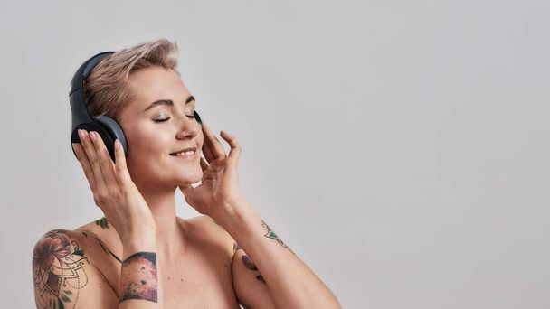 Clear Sound. Attractive tattooed woman with pierced nose and short hair in headphones enjoying listening to music isolated over grey background - Photo, image