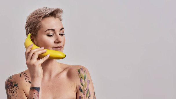 Eat Fruits. Attractive tattooed woman with pierced nose and short hair holding banana as a phone isolated over grey background - Photo, Image