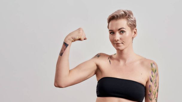 Eu sou forte. Retrato de mulher tatuada semi-nua com cabelo curto olhando para a câmera, mostrando seu músculo forte, bíceps isolado sobre fundo claro
 - Foto, Imagem