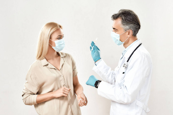 Preventing virus. Professional male doctor wearing protective mask and sterile gloves showing syringe with vaccine to a young female patient - Zdjęcie, obraz