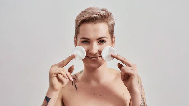 Πορτρέτο της όμορφης γυναίκας με τατουάζ με τρυπημένη μύτη και κοντά μαλλιά χαμογελώντας ενώ κρατώντας βαμβάκι μαξιλάρια για την αφαίρεση μακιγιάζ, τον καθαρισμό του δέρματος απομονώνονται σε γκρι φόντο - Φωτογραφία, εικόνα