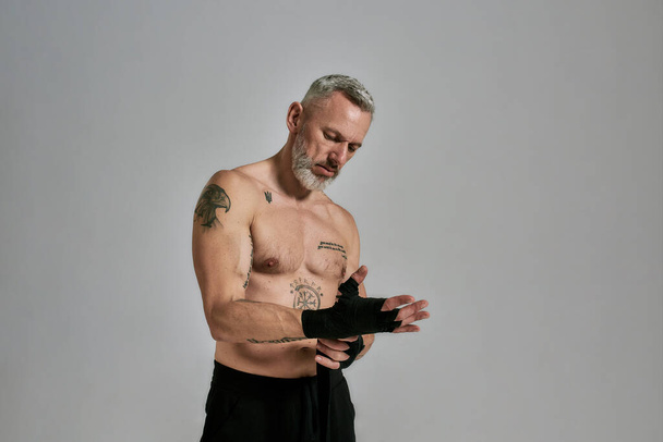 Half naked middle aged athletic man, kickboxer wrapping hands for Muay Thai, Boxing or Kickboxing, standing in studio over grey background - Foto, Bild
