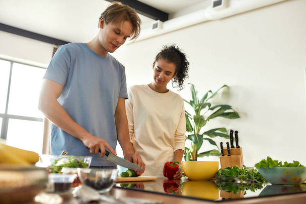Young man cutting vegetables while woman holding pepper and watching him. Vegetarians preparing healthy meal in the kitchen together. Vegetarianism, healthy food concept - Photo, Image