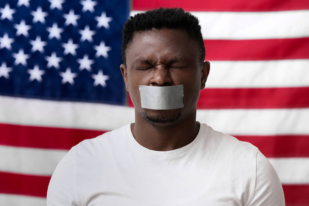 Trauriger afroamerikanischer Mann mit verklebtem Mund in der Nähe der Nationalflagge der USA. Schluss mit Rassismus - Foto, Bild