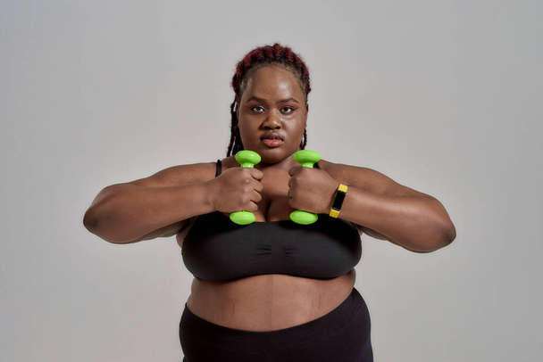 Changes. Plump, plus size african american woman in sportswear exercising, holding green dumbbells, posing in studio over grey background - 写真・画像