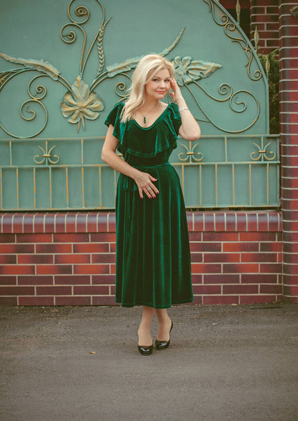 Žena středního věku v klasických zelených sametových šatech a smaragdových doplňcích na procházku v malém domku. Koncept retro a retro ženskosti - Fotografie, Obrázek