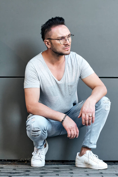 a stylish guy with glasses sat down near a gray wall and looks away. strong and strict male gaze. light jeans and a fashionable gray T-shirt. sunlight. half-length portrait - Photo, Image