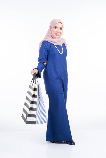 Ένα όμορφο μουσουλμανικό θηλυκό μοντέλο σε ένα ασιατικό παραδοσιακό φόρεμα μεταφέρουν τσάντες ψώνια που απομονώνονται σε λευκό φόντο. Idul fitri εορταστική προετοιμασία έννοια για ψώνια. Πλήρες μήκος πορτρέτο. - Φωτογραφία, εικόνα