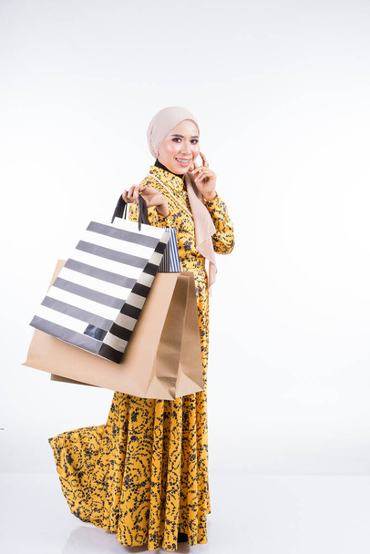 Egy gyönyörű muszlim női modell ázsiai hagyományos ruhában, fehér háttérrel elszigetelt bevásárlószatyrokkal. Idul fitri ünnepi előkészületi vásárlási koncepció. Teljes hosszúságú portré. - Fotó, kép
