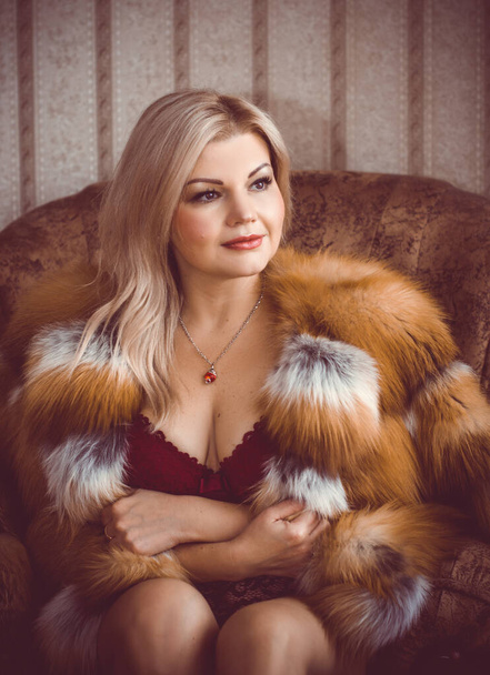 Mulher sexy de meia idade em corpo de lingerie vermelha e casaco de raposa de pele, em ambiente acolhedor em casa na noite quente de inverno. Conceito de feminilidade e ternura após 30-40 anos
 - Foto, Imagem