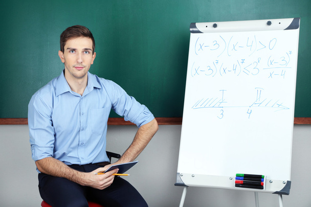 νεαρός δάσκαλος κάθεται κοντά chalkboard σε σχολική τάξη - Φωτογραφία, εικόνα