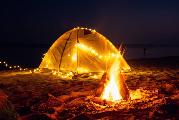 The fire at night on the beach. Summer mood. Night camping with a retro garland on sea shore. - Photo, Image
