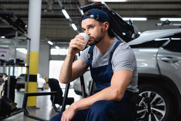 mechanic in cap drinking coffee to go near car in workshop - Photo, Image