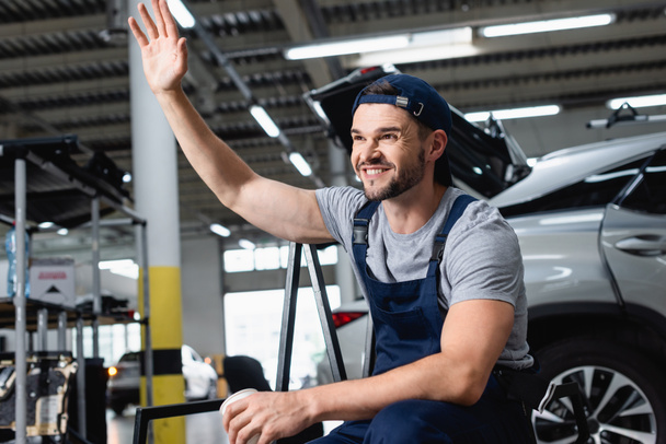 happy mechanic in cap waving hand and holding paper cup in workshop - Photo, Image