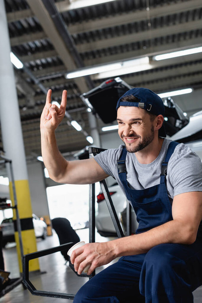 Glücklicher Mechaniker mit Mütze zeigt Felsschild und hält Pappbecher in Autowerkstatt - Foto, Bild