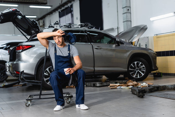 tired technician holding cap and paper cup while sitting near cars in workshop  - Photo, Image