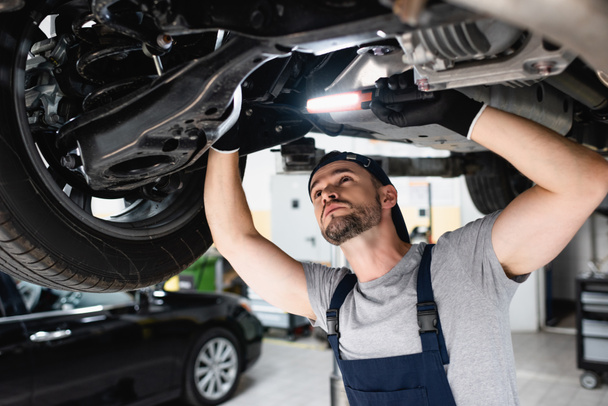 handsome mechanic holding flashlight and fixing car in service station  - Photo, Image