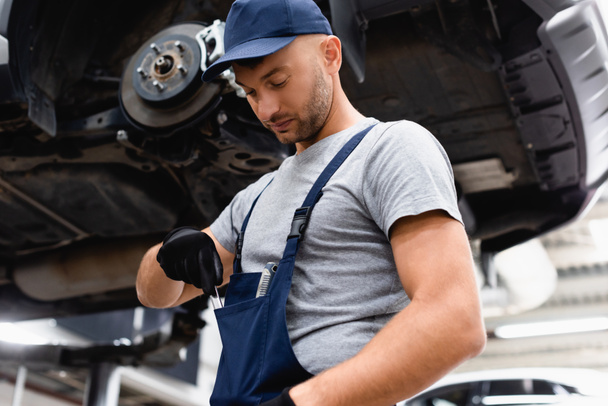 low angle view of mechanic in overalls taking wrench from pocket near car - Fotografie, Obrázek