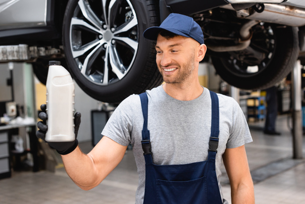 selective focus of happy mechanic in cap and overalls holding bottle with car oil  - Foto, afbeelding