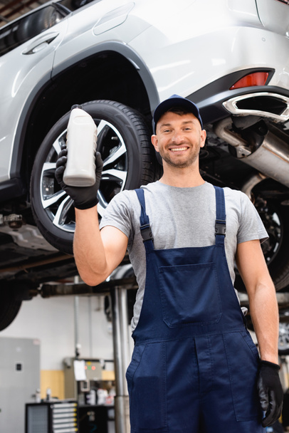 low angle view of cheerful mechanic in cap holding bottle with car oil  - Photo, Image